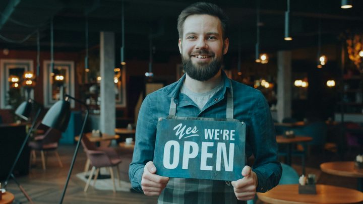 A smiling shop owner in an apron holding a Yes We're Open sign in his restaurant